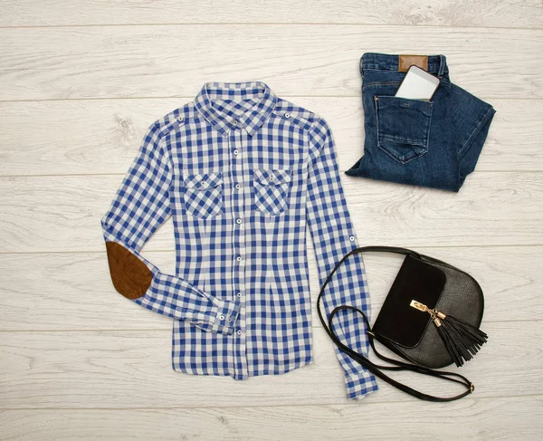 Mavi ve beyaz ekose gömlek, kot pantolon, bir telefon, siyah bir çanta. Ahşap arka plan. Moda kavramı, üstten görünüm — Stok fotoğraf