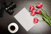 Čistý list papíru, růžové tulipány, kamery a hrnek kávy. Černý stůl. pohled shora