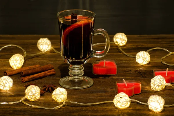 Κούπα ζεστό κρασί με μπαχαρικά, κεριά σε σχήμα καρδιάς σε ένα ξύλινο τραπέζι, μια γιρλάντα από φανάρια. Ξυλάκια κανέλας, γλυκάνισο — Φωτογραφία Αρχείου