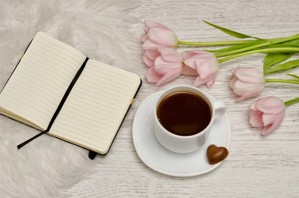Taza de café con caramelos, un cuaderno con páginas en blanco y tulipanes rosados sobre la mesa . — Foto de Stock