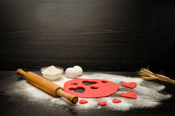 Massa de farinha vermelha com corações esculpidos em uma mesa preta. Rolo de pino, farinha, ovos e orelhas de trigo. Lugar para texto — Fotografia de Stock