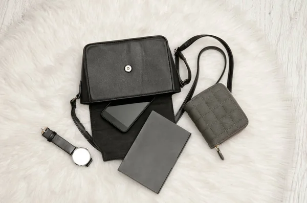 Open zwarte tas met decoratieve dingen, laptop, mobiele telefoon, horloge en grijs portemonnee. De witte vacht op achtergrond, bovenaanzicht. Fashion concept — Stockfoto