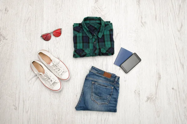Mavi-yeşil kareli gömlek, gözlük, Ayakkabı, kot pantolon, telefon ve pasaport. Ahşap arka plan. Moda kavramı, üstten görünüm — Stok fotoğraf