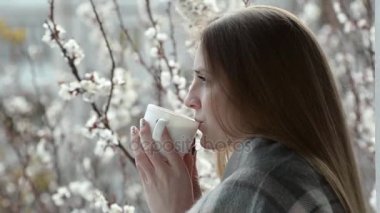 Bir balkon cam kenarında duran ve çiçek açan kayısı bir arka plan üzerinde çay içme bir ekose kız