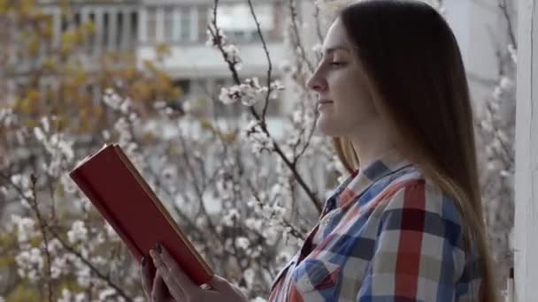 Девушка в клетчатой рубашке стоит на балконе у окна и читает книгу на фоне цветущих абрикосов — стоковое видео