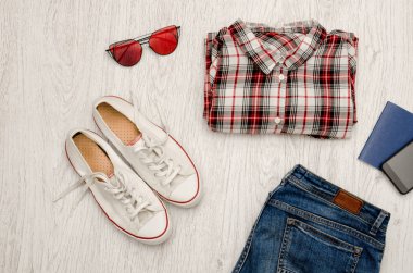 Kırmızı ve beyaz kareli gömlek, gözlük, Ayakkabı, kot pantolon, telefon ve pasaport. Ahşap arka plan. Moda kavramı, üstten görünüm