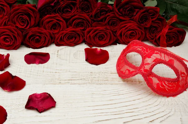 Máscara de carnaval vermelho encontra-se em uma mesa de madeira branca no fundo de um buquê de rosas — Fotografia de Stock