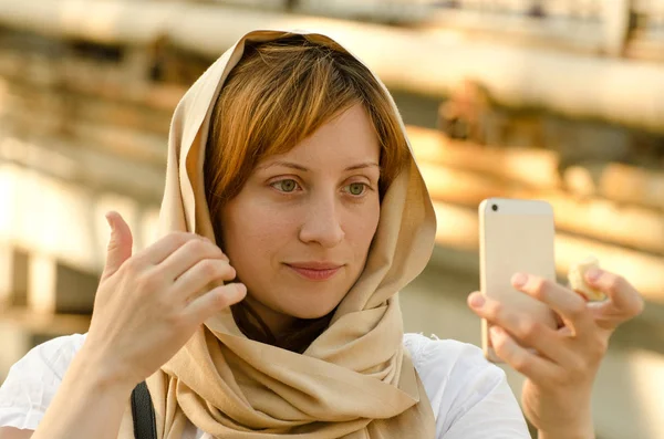 Beautiful red-haired girl in a scarf looks in a smartphone — Stock Photo, Image