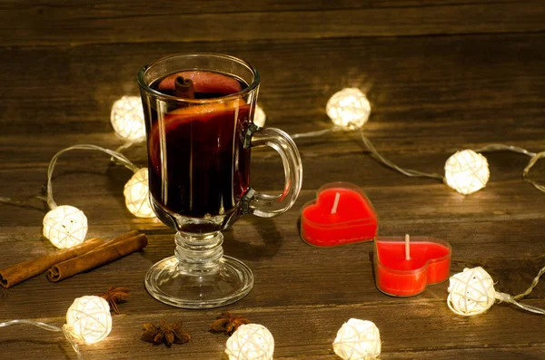 Κούπα ζεστό κρασί με μπαχαρικά, κεριά σε σχήμα καρδιάς σε ένα ξύλινο τραπέζι, μια γιρλάντα από φανάρια. Ξυλάκια κανέλας, γλυκάνισο — Φωτογραφία Αρχείου