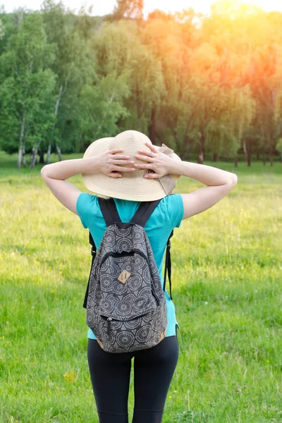 Güneşli bir sırt çantası ayakta karşı bir yeşil orman ile şapkalı kız. Arkadan Görünüm
