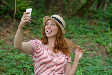 Şapkalı Kızıl saçlı kız komik selfie bir yeşil orman arka plan üzerinde yapar.