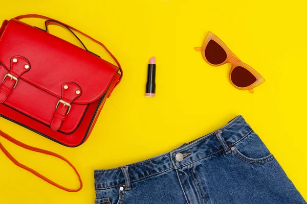 Kırmızı el çantası, kot şort, güneş gözlüğü ve ruj. Parlak sarı arka plan. Moda kavramı