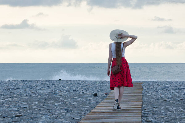 Girl in a red skirt and hat walks on the deck on the beach against the backdrop of the sea and sky. Back view