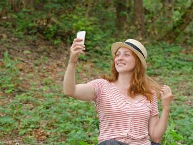 Şapkalı Kızıl saçlı kız selfie yeşil orman bir arka plan üzerinde yapar.