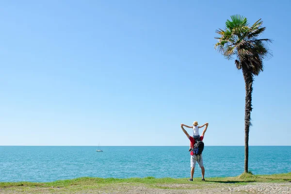 Pai com filho sobre ombros fica para trás contra o pano de fundo de palma alta, mar e céu azul — Fotografia de Stock