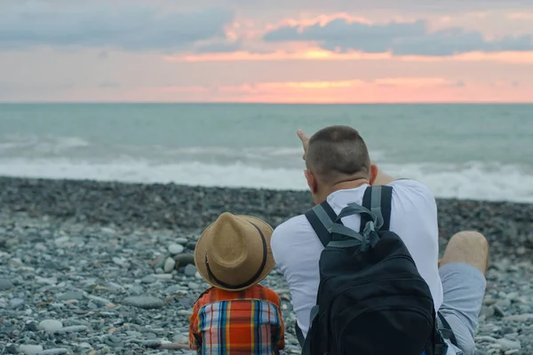 Genç Baba ve oğul deniz ve güneşin zemin karşı sahilde oturuyorlar.