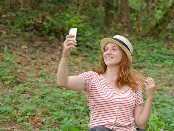Şapkalı Kızıl saçlı kız selfie yeşil orman bir arka plan üzerinde yapar.
