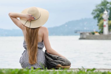 İskelede oturup bir sırt çantası ile şapkalı kız. Dağlar ve deniz feneri arka planda. Arkadan görüntülemek