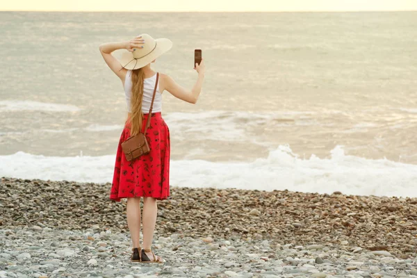 Chica en falda roja y sombrero haciendo selfie en la playa en el fondo del mar y el cielo. Vista desde atrás — Foto de Stock