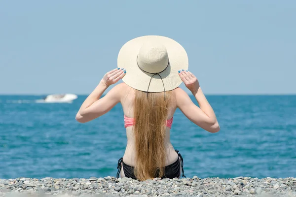 Красива дівчина в капелюсі і купальник сидить на пляжі. Море і човен на фоні. Вид ззаду — стокове фото