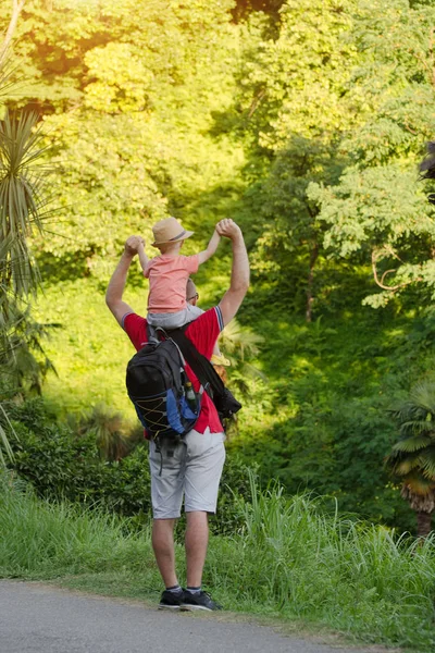 Junger Vater mit Sohn auf den Schultern, der vor dem Hintergrund tropisch grünen Waldes steht. Rückseite. Sonnenlicht. — Stockfoto