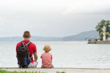 Baba ve oğul deniz arka plan, deniz feneri ve dağlar uzakta iskelede oturup. Arkadan Görünüm