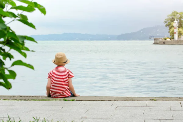 Αγόρι σε ένα καπέλο και ριγέ μπλουζάκι που κάθεται στην παραλία. Θέα από το πίσω μέρος — Φωτογραφία Αρχείου