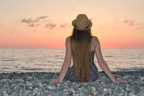 Deniz kıyısı üzerinde oturan şapkalı kız. Günbatımı zamanı. Arkadan görüntülemek