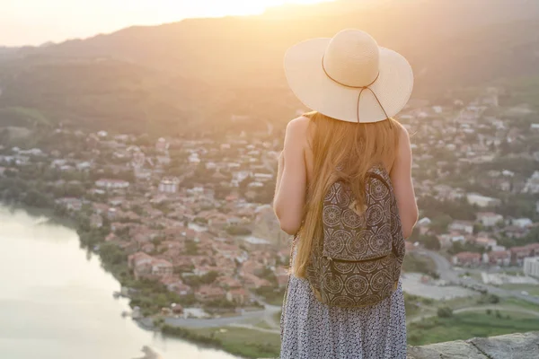 Beautiful girl with a backpack and in a hat looks at the city and the river below. Back view — Stock Photo, Image