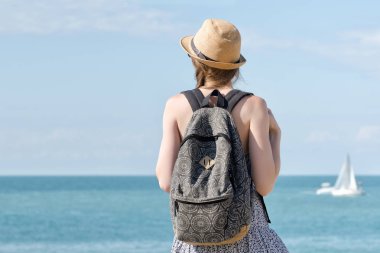Bir sırt çantası konumu sahil şeridi üzerinde olan şapkalı kız. Yelkenli uzakta. Arkadan görüntülemek