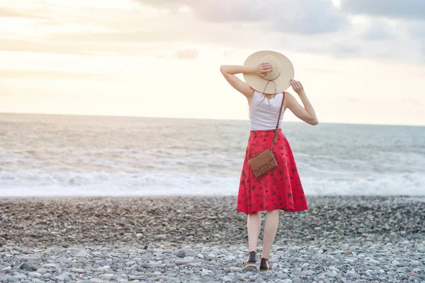 Menina em uma saia vermelha e chapéu está em pé junto ao mar, pôr do sol. Vista de trás — Fotografia de Stock