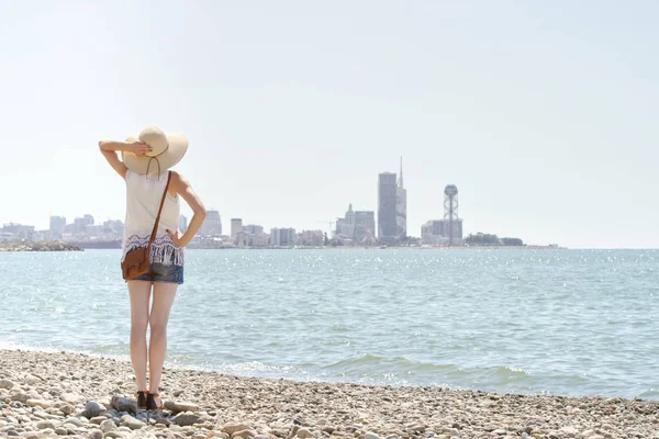 Chica con un sombrero de pie en la orilla del mar. Ciudad en la distancia. Vista desde atrás — Foto de Stock