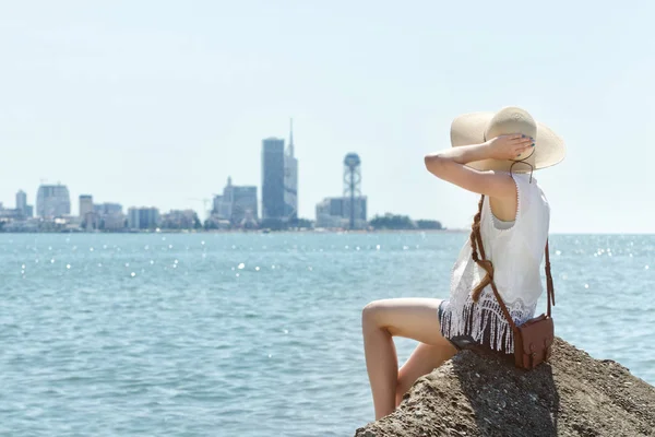 Deniz kenarında bir kayaya oturan şapkalı kız. Mesafe şehirde. Güneşli gün