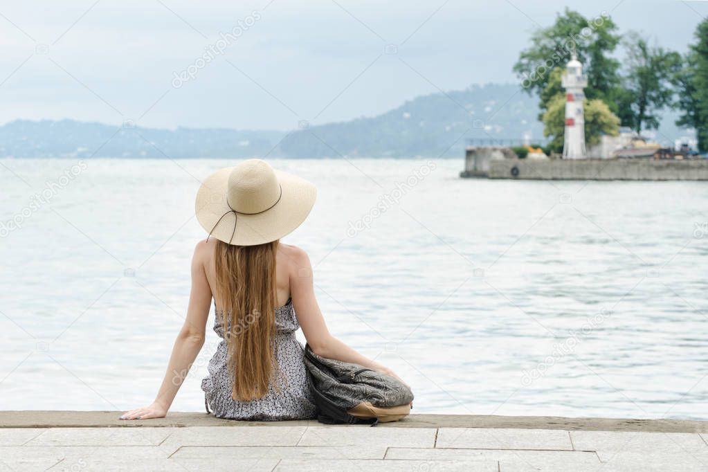 Girl in a hat with a backpack sitting on the pier. Mountains and lighthouse on the background. View from the back