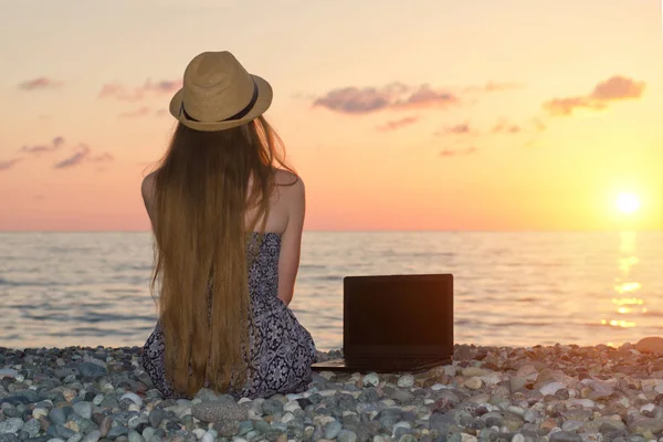 Κορίτσι σε ένα καπέλο κάθεται στην παραλία, κοντά σε ένα φορητό υπολογιστή. Θάλασσα στο ηλιοβασίλεμα — Φωτογραφία Αρχείου