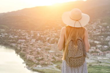 Güzel kız bir şapka ve bir sırt çantası ile şehir ve nehir aşağı bakar. Arkadan Görünüm