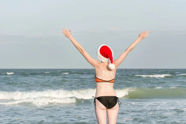 Dziewczyna w Santa kapelusze z napisem nowy rok z tyłu. Brzeg morza. Podniósł ręce. Widok z tyłu — Zdjęcie stockowe