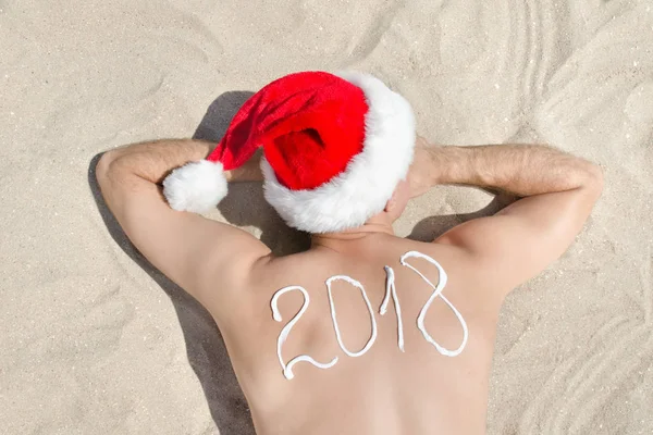 Mann mit Weihnachtsmannmütze mit der Aufschrift 2018 auf dem Rücken im Sand liegend. Blick von oben — Stockfoto