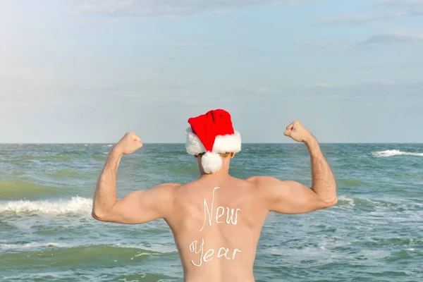 Strong man in Santa hats with the inscription New Year on the back standing on the beach. Back view — Stock Photo, Image