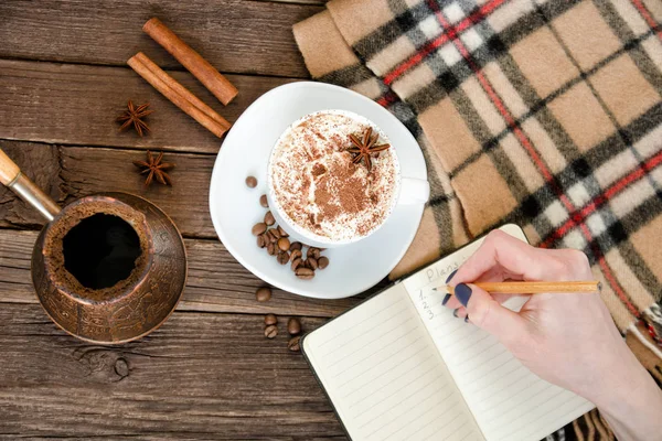 Bir kafede planlama. Kupa latte, içinde a defter bir kalem ile yazılı bir el. Üstten Görünüm