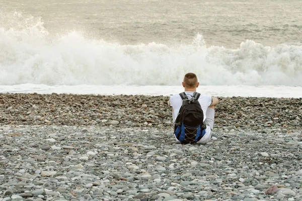 Genç adam bir sırt çantası ile okyanus kıyısında oturuyor. Arkadan görüntülemek — Stok fotoğraf