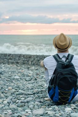 Adam şapka ve zemin deniz ve gün batımı gökyüzünün karşı sahilde oturup sırt çantası ile. Arkadan Görünüm