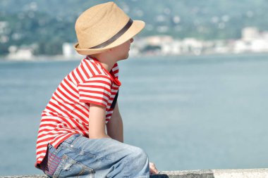 Çocuk bir şapka ile sahilde oturuyor ve çizgili tişört, mesafe içine görünüyor. Yan görünüm
