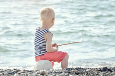 Boy Çizgili tişört içinde bir değnek ile sahilde oturuyor. Deniz içinde belgili tanımlık geçmiş