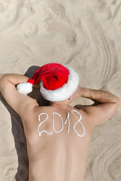 Mann mit Weihnachtsmannmütze mit der Aufschrift 2018 auf dem Rücken im Sand liegend. Blick von oben — Stockfoto