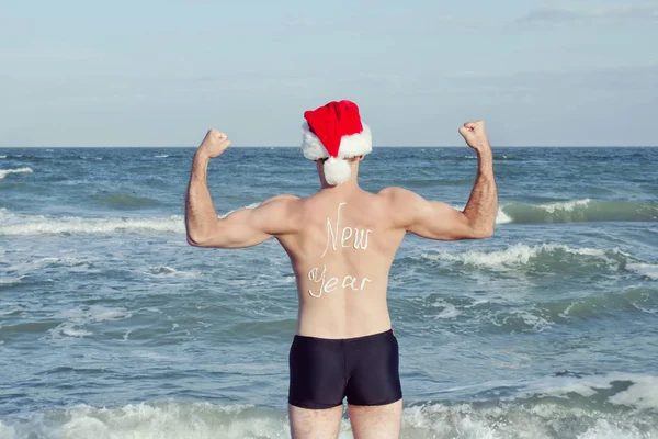 Starker Mann in Weihnachtsmannmützen mit der Aufschrift Neujahr auf dem Rücken, der am Strand steht. zurück — Stockfoto