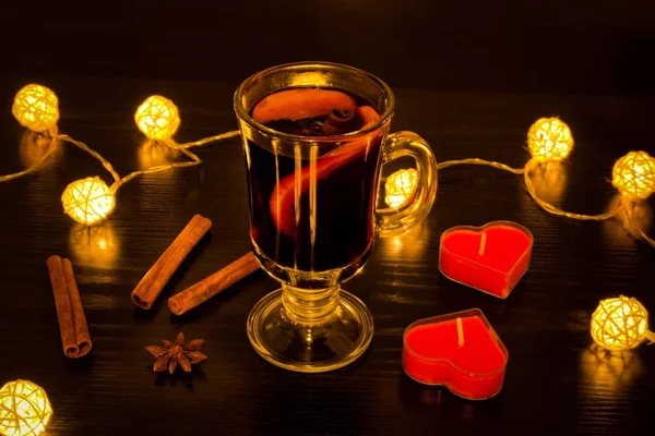 Mug of mulled wine with spices, candles in the shape of a heart, cinnamon sticks, star anise. Illumination of rattan lanterns on a black wooden table — Stock Photo, Image