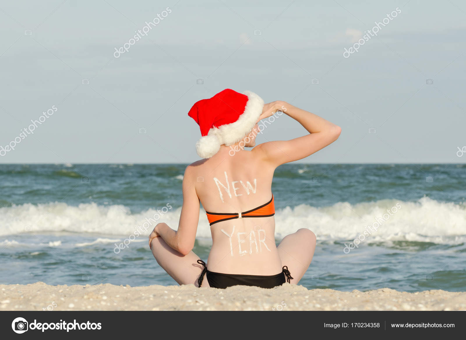 背中に碑文の新年とサンタ帽子の女の子 ビーチに座っているし 遠くに見える ストック写真 C Somemeans