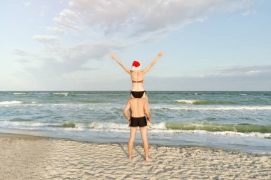 Noel Baba şapkalı kız bir adam omuzlarında oturur. Deniz kıyısında. Arkadan Görünüm