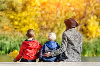 Anne ve iki çocuğu iskelede oturuyor. Güneşli sonbahar. Arkadan Görünüm